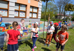 Pięknie świeci słońe, widok na dwie grupy dzieci malujących na foli piękne kolorowe obrazy,w tle drzewa oraz pomarańczowy budynek przedszkola